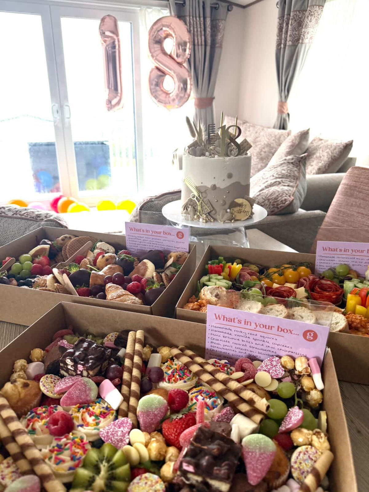 Dessert, savoury and brunch boxes with colourful treats on a table, with balloons and a cake in the background.