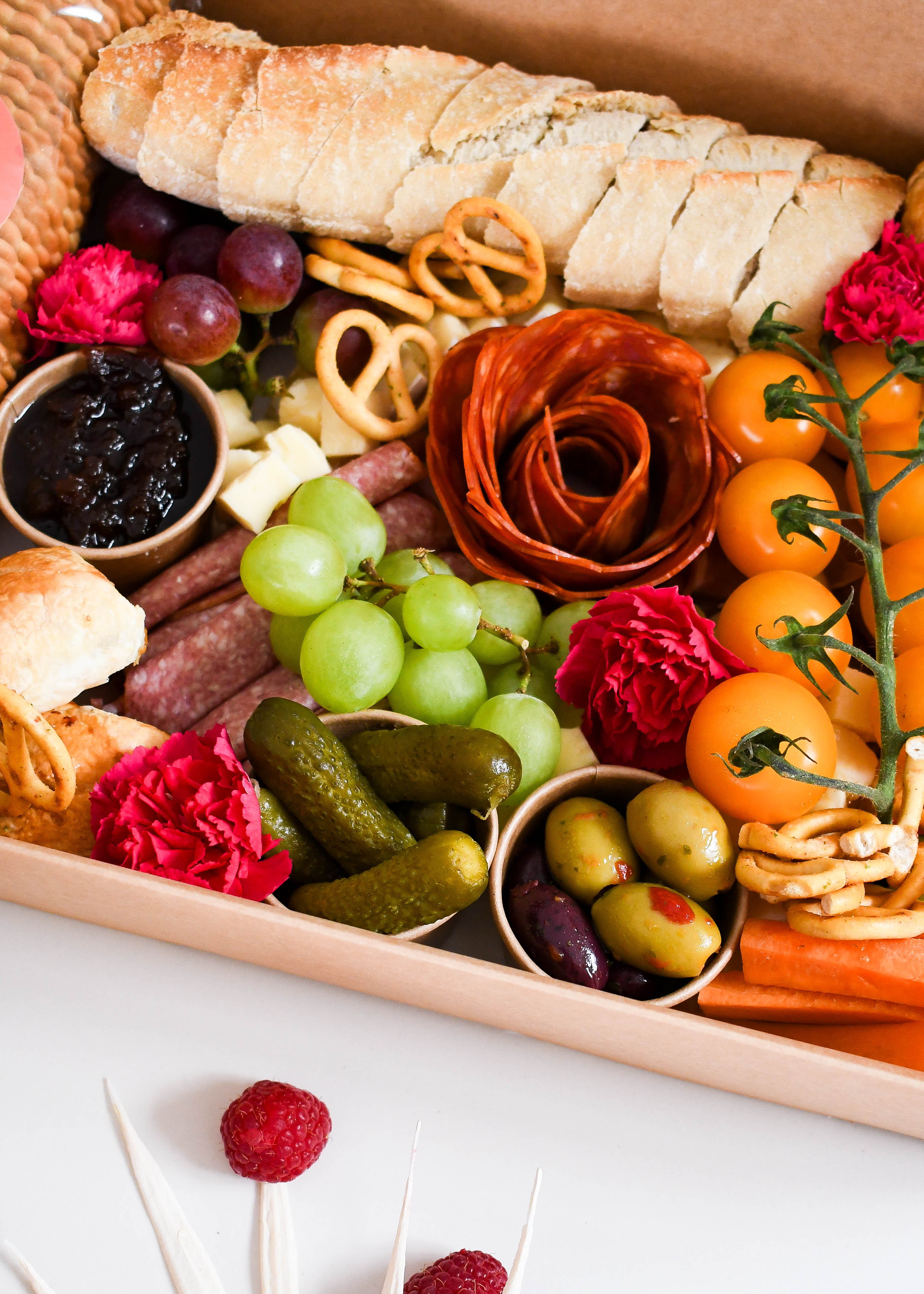 Assorted food platter with bread, fruits, vegetables, and flowers on a white surface.