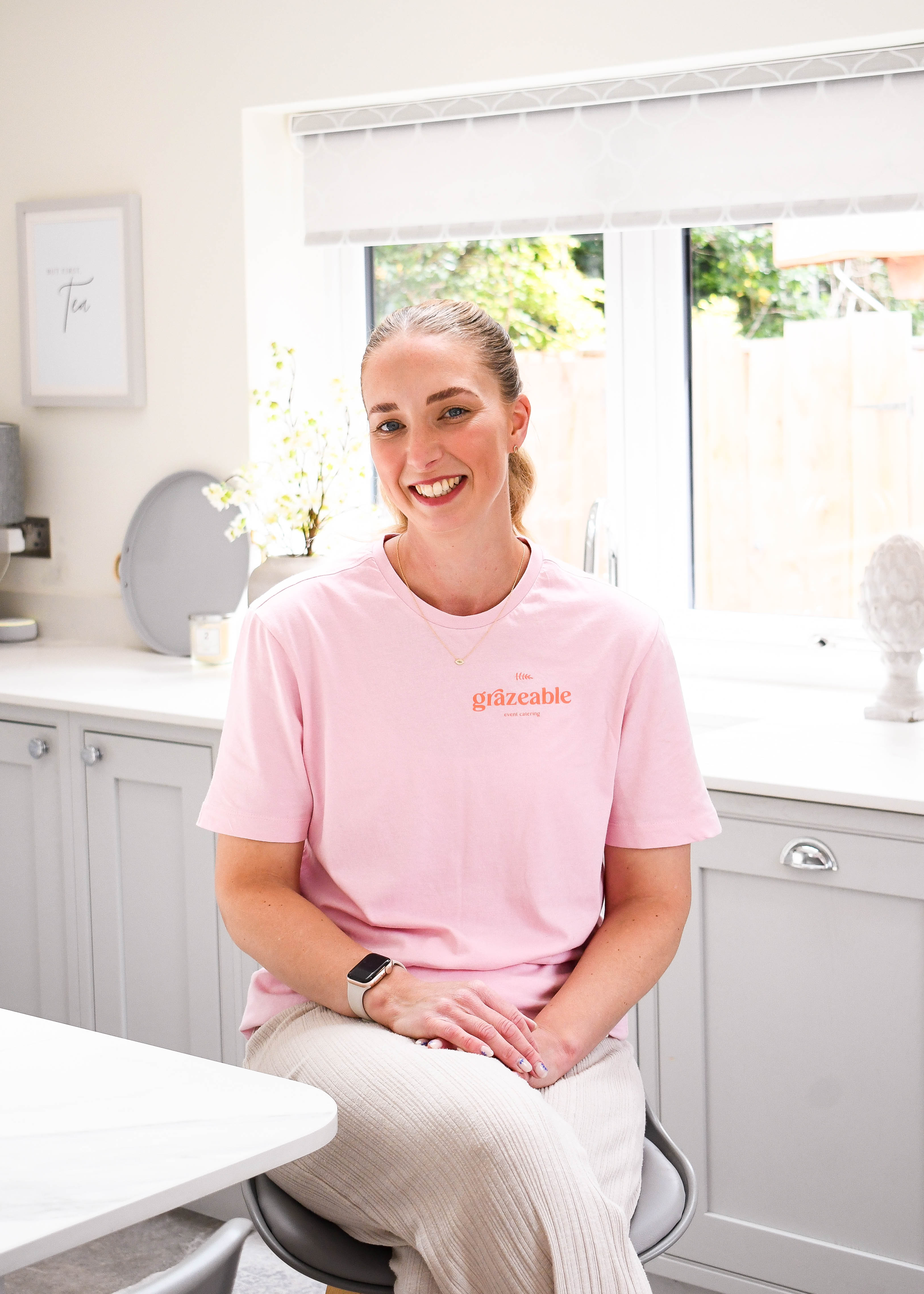 Woman wearing a pink t-shirt with 'grazeable' text, sitting in a kitchen.
