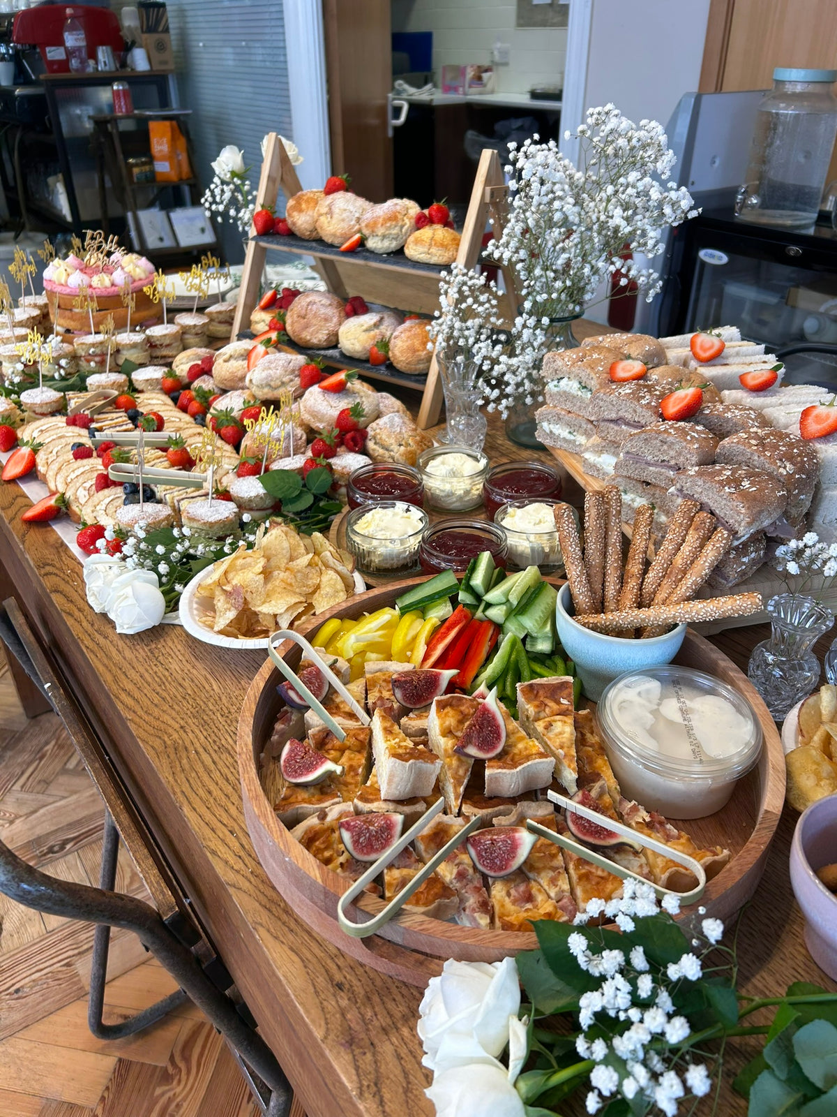 Diverse array of food items on a wooden table with flowers and a cafe background.