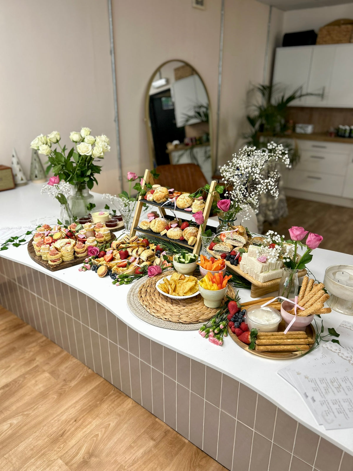 Styled afternoon tea table with wooden platters and risers styled with flowers on a white countertop surface