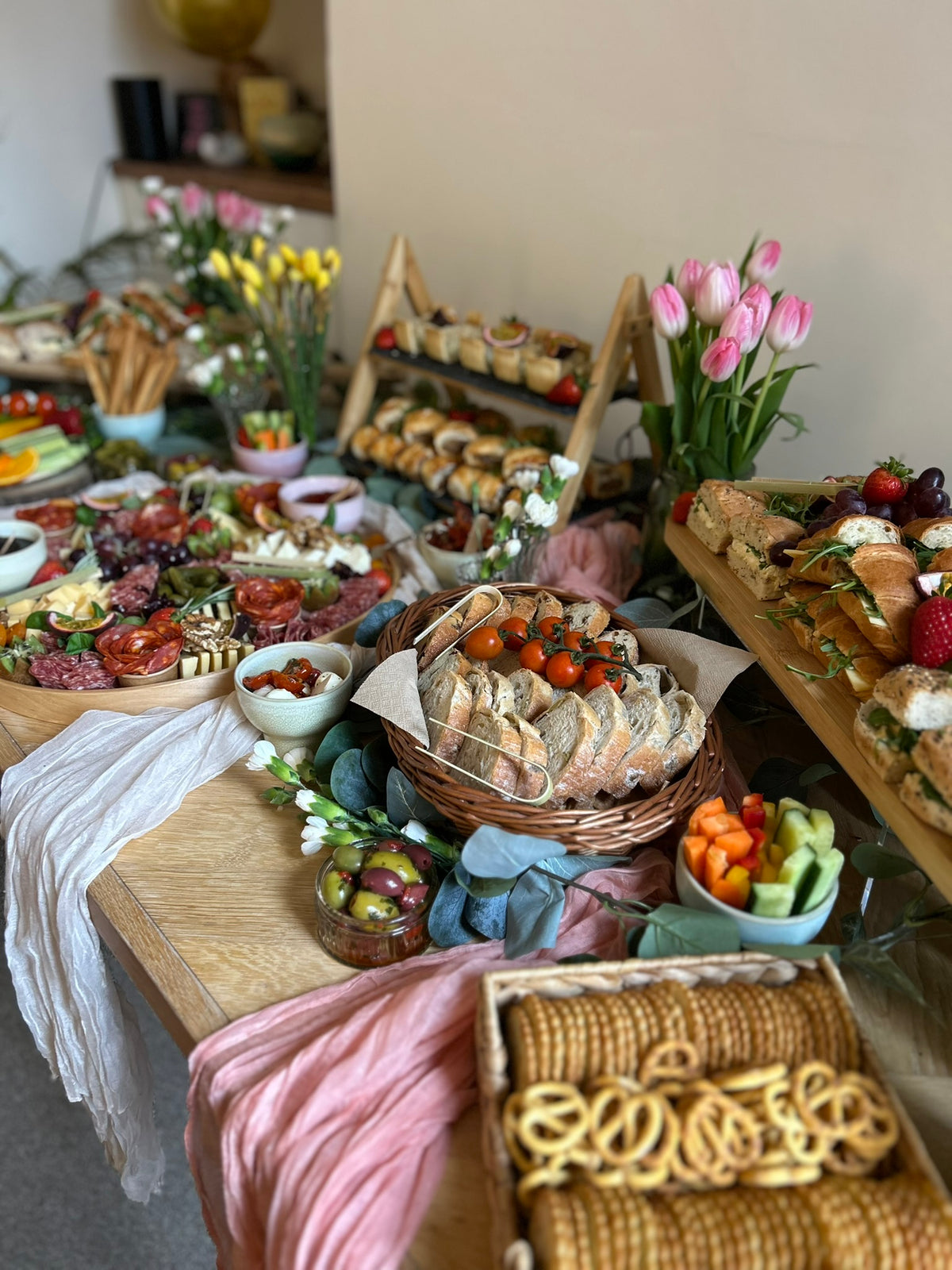 Styled grazing table with pastel cloths and wooden risers with various food items including bread, fruits, and vegetables in a home setting.